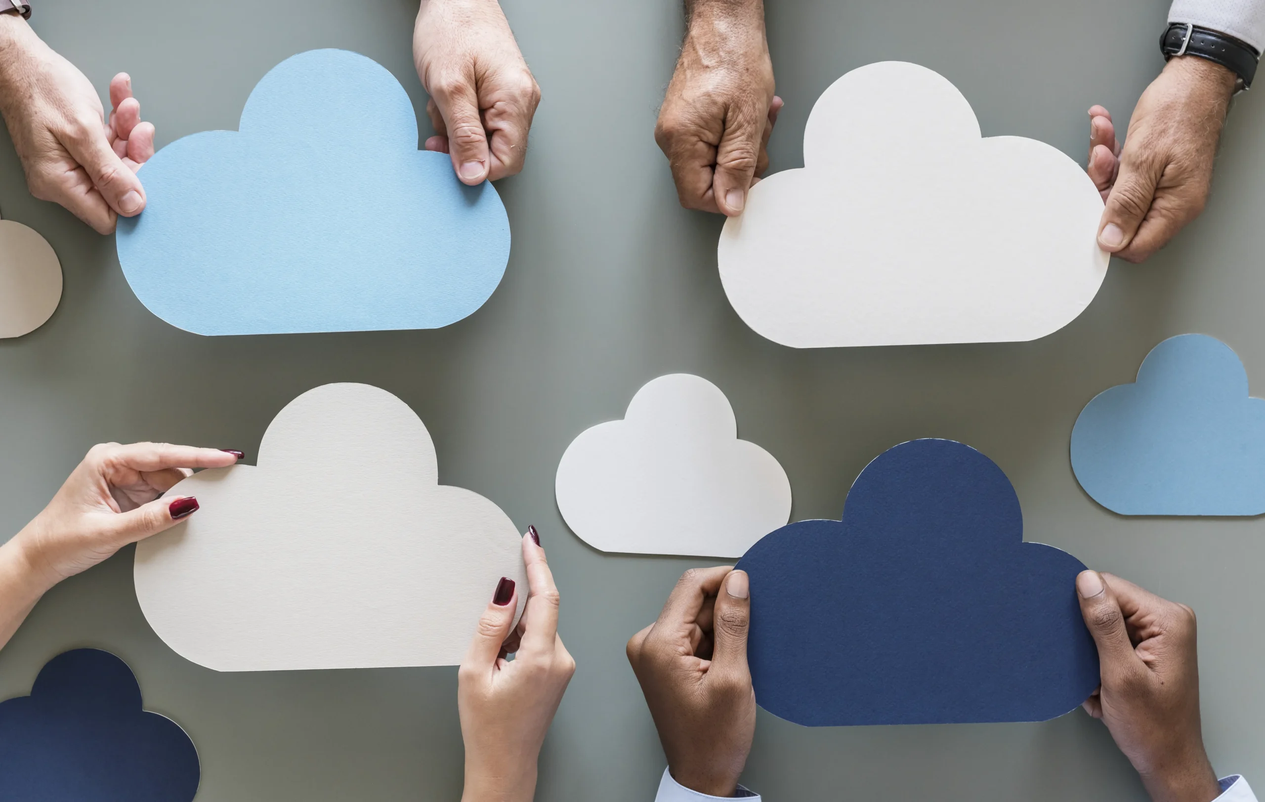 Dažādu krāsu papīra mākoņi cilvēku rokās – simbolizē sadarbību mākoņpakalpojumu pārvaldībā un dažādu platformu (Azure, AWS, Google Cloud) integrāciju